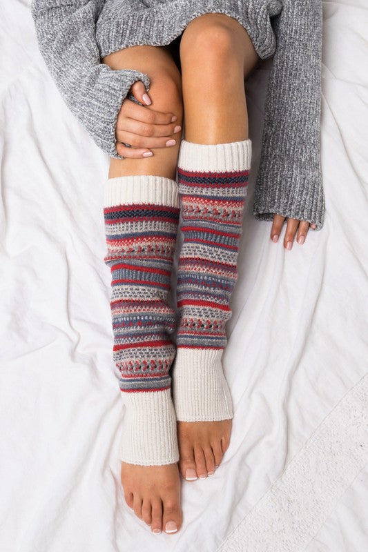 A person wearing a grey sweater and the Short Fairisle Legwarmer sits on a white bed, legs extended and hands resting gently on the soft spun acrylic.