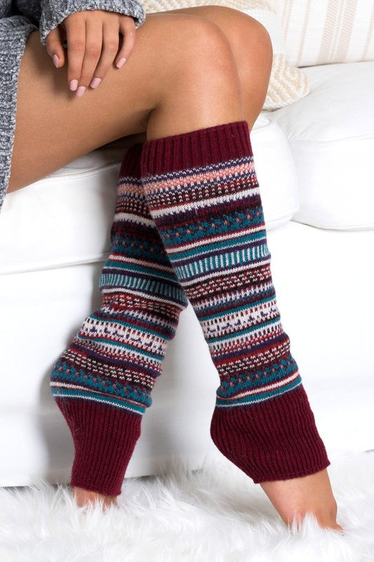 A person wearing a grey sweater and the Short Fairisle Legwarmer sits on a white bed, legs extended and hands resting gently on the soft spun acrylic.