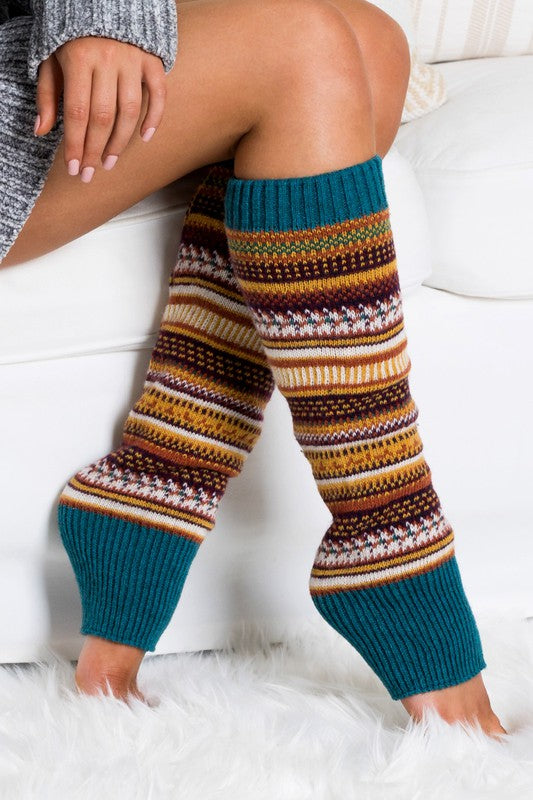 A person wearing a grey sweater and the Short Fairisle Legwarmer sits on a white bed, legs extended and hands resting gently on the soft spun acrylic.