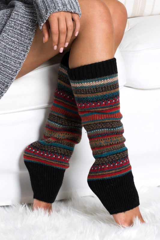 A person wearing a grey sweater and the Short Fairisle Legwarmer sits on a white bed, legs extended and hands resting gently on the soft spun acrylic.