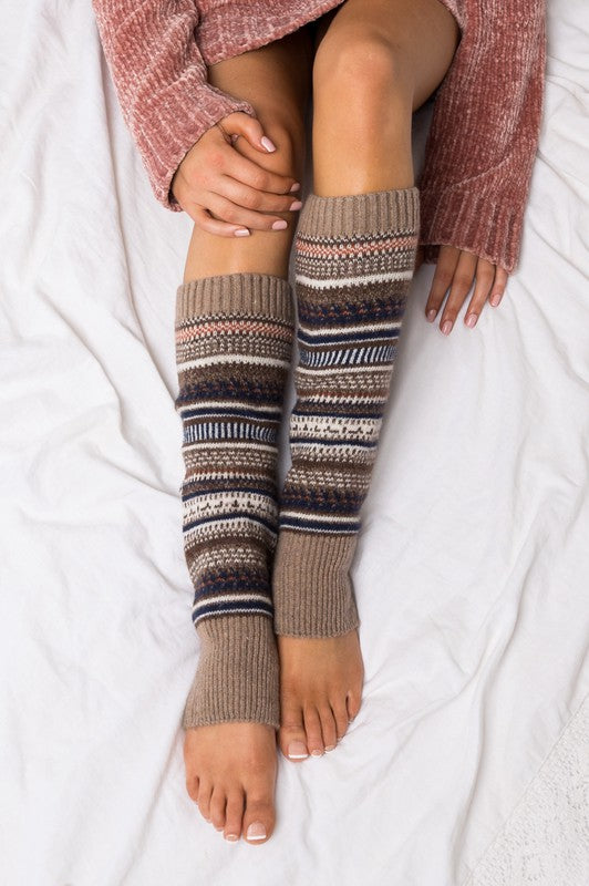 A person wearing a grey sweater and the Short Fairisle Legwarmer sits on a white bed, legs extended and hands resting gently on the soft spun acrylic.