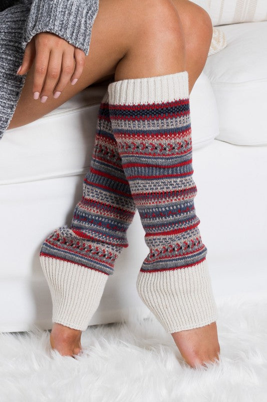 A person wearing a grey sweater and the Short Fairisle Legwarmer sits on a white bed, legs extended and hands resting gently on the soft spun acrylic.