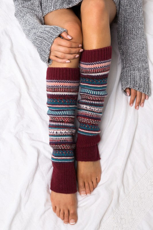 A person wearing a grey sweater and the Short Fairisle Legwarmer sits on a white bed, legs extended and hands resting gently on the soft spun acrylic.