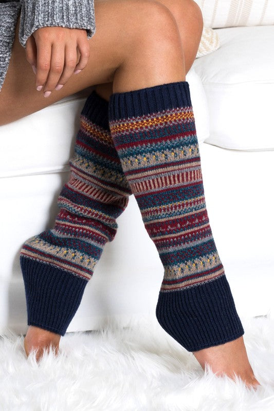 A person wearing a grey sweater and the Short Fairisle Legwarmer sits on a white bed, legs extended and hands resting gently on the soft spun acrylic.