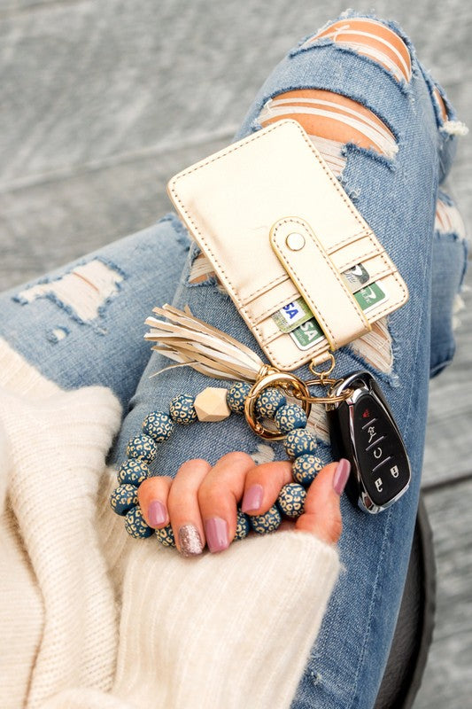 Woman holding a Leopard Beaded Key Ring Wallet Bracelet with a beige tassel and laser cut leopard beads, attached to a white wallet, against a gray sweater and blue ripped jeans.