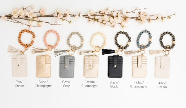 Woman holding a Leopard Beaded Key Ring Wallet Bracelet with a beige tassel and laser cut leopard beads, attached to a white wallet, against a gray sweater and blue ripped jeans.