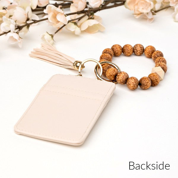 Woman holding a Leopard Beaded Key Ring Wallet Bracelet with a beige tassel and laser cut leopard beads, attached to a white wallet, against a gray sweater and blue ripped jeans.