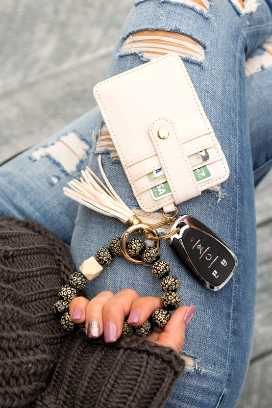 Woman holding a Leopard Beaded Key Ring Wallet Bracelet with a beige tassel and laser cut leopard beads, attached to a white wallet, against a gray sweater and blue ripped jeans.