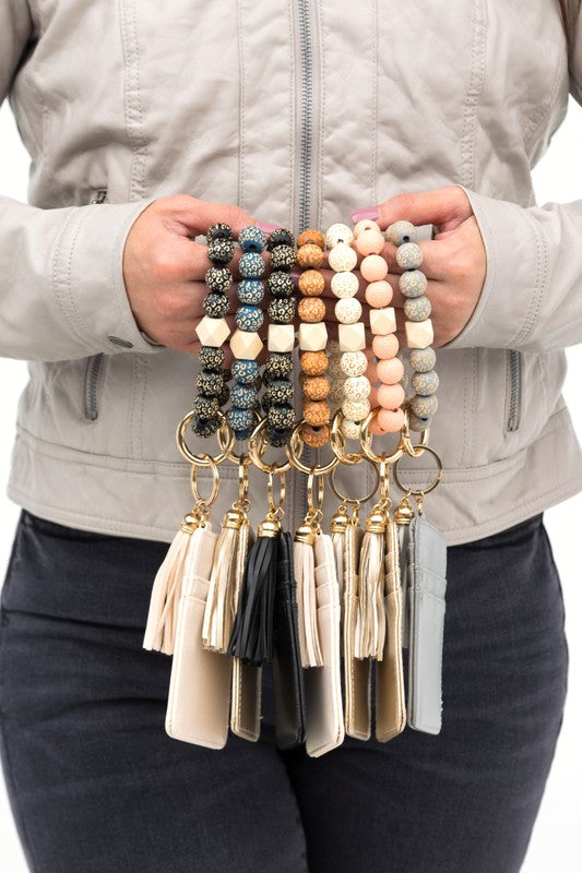 Woman holding a Leopard Beaded Key Ring Wallet Bracelet with a beige tassel and laser cut leopard beads, attached to a white wallet, against a gray sweater and blue ripped jeans.