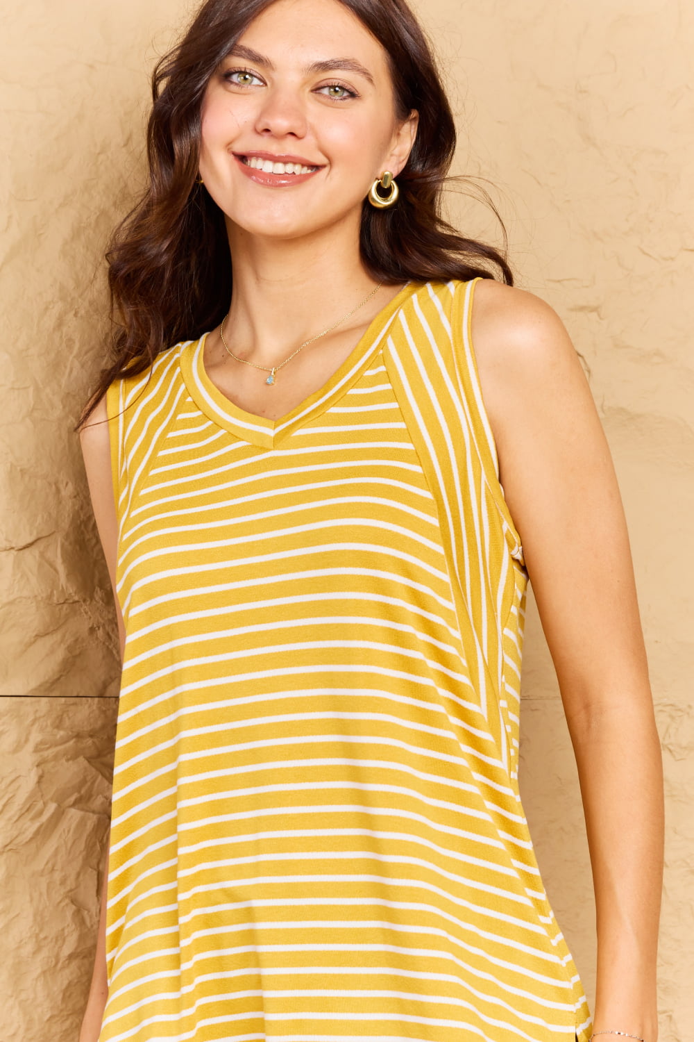 A woman wearing the timeless Doublju Talk To Me Full Size Striped Sleeveless V-Neck Top in yellow and white, paired with distressed denim shorts, poses against a textured beige wall.