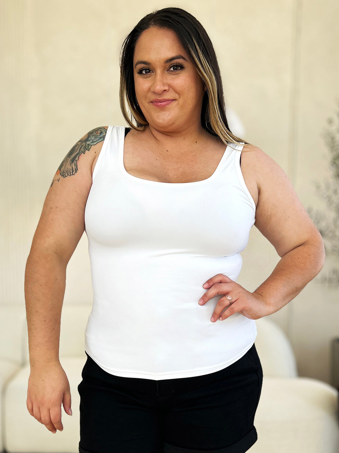 A woman with brown hair, wearing sunglasses, blue jeans, and the Basic Bae Full Size Square Neck Wide Strap Tank in black stands against a concrete wall, showcasing a basic style.