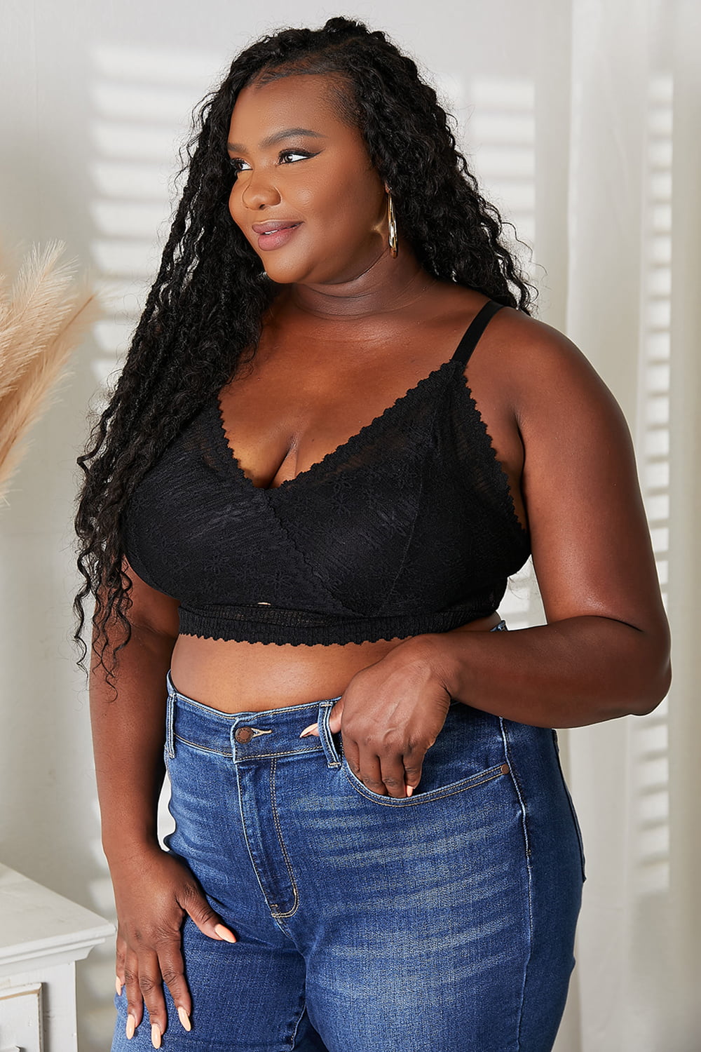 A woman with long black braids is smiling while wearing the JadyK Skye Full Size Lace Bralette and blue jeans, standing near a plant in a white pot.