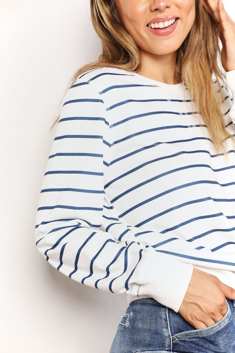 A person smiling while wearing the Double Take Striped Long Sleeve Round Neck Top, paired with denim shorts, stands in front of a white background with a plant.