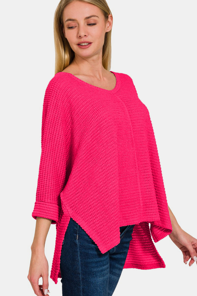 A woman wearing the Zenana V-Neck High-Low Jacquard Knit Top in a bright pink, loose-knit style with three-quarter sleeves, paired with blue jeans, stands against a plain white background, showcasing her comfortable knit top.