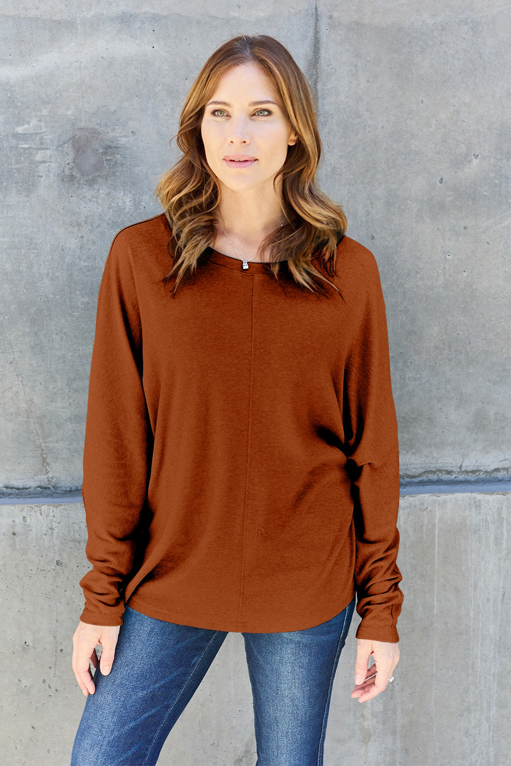 A person wearing a Double Take Full Size Round Neck Long Sleeve T-Shirt in green, blue distressed jeans with a slightly stretchy fit, and a crossbody bag, holding a beverage in a plastic cup.