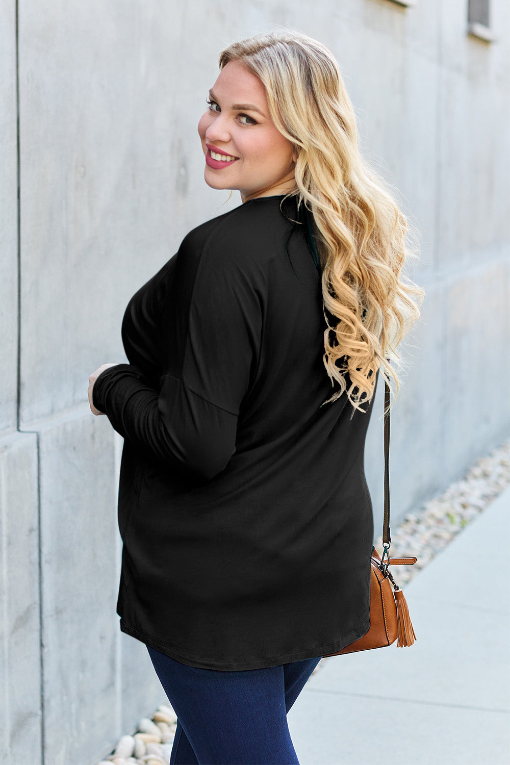 A woman with shoulder-length hair wearing a Basic Bae Full Size Round Neck Dropped Shoulder T-Shirt in green and a pair of jeans, exuding a basic style, stands against a concrete wall. She's holding an imported brown woven backpack on her shoulder while touching her hair with her hand.