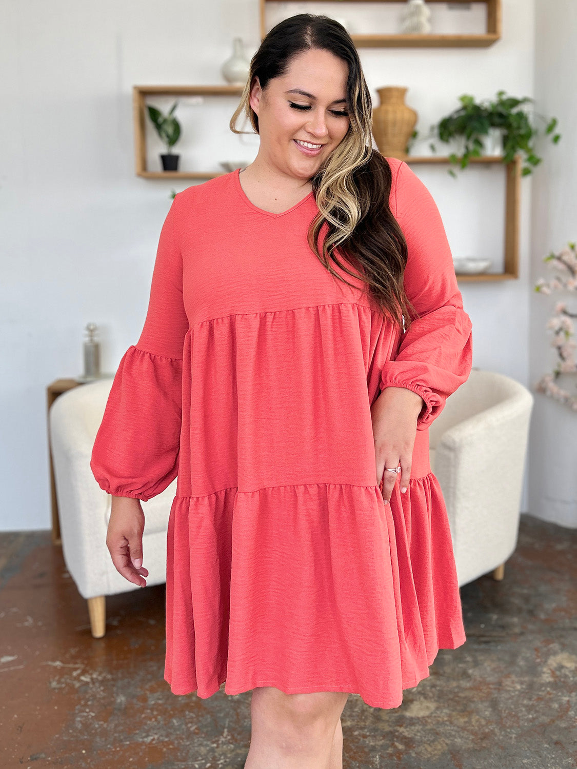 A woman stands wearing the Double Take Full Size V-Neck Balloon Sleeve Tiered Dress with Pockets in pink. Crafted from high stretch yarn, this dress combines comfort and style effortlessly. She has long, wavy hair and accessorizes with hoop earrings and a necklace. This imported garment is easy to care for – simply machine wash cold to keep it looking fresh.