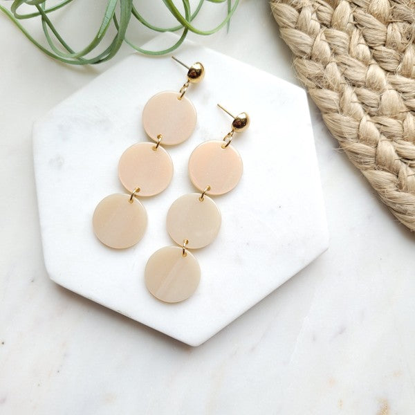 The Elle Earrings - Linen are a pair of dangling earrings featuring three circular beige pendants on each side, crafted from lightweight acrylic with a subtle rose gold shimmer, artfully displayed on a white rectangular dish set on a wooden surface.