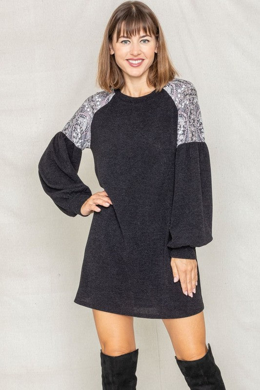 A person with long blonde hair smiles while wearing a dark Paisley Shoulder Accent Tunic Dress, featuring delicate lace detail on the shoulders. Made in the United States, this dress offers added comfort with its blend of 95% polyester and 5% spandex.