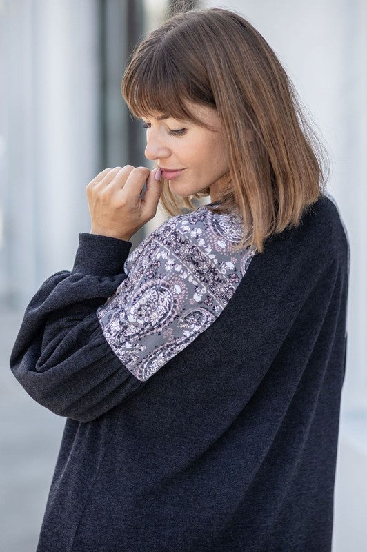 A person with long blonde hair smiles while wearing a dark Paisley Shoulder Accent Tunic Dress, featuring delicate lace detail on the shoulders. Made in the United States, this dress offers added comfort with its blend of 95% polyester and 5% spandex.