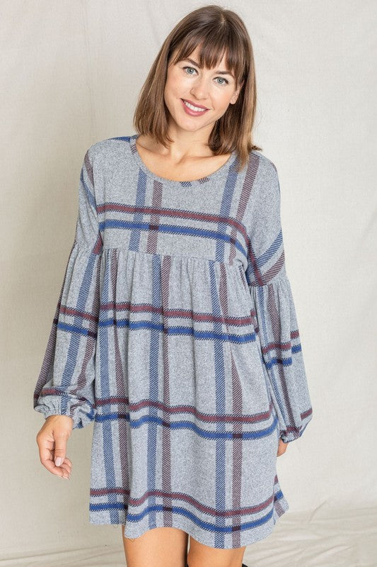 A person with curly hair wearing a Plus Plaid Empire Waist Balloon Sleeve Mini Dress stands against a white wall, smiling.