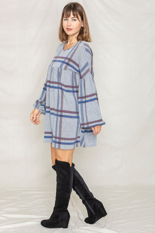 A person with curly hair wearing a Plus Plaid Empire Waist Balloon Sleeve Mini Dress stands against a white wall, smiling.