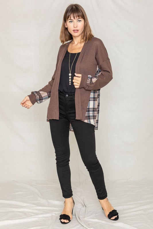 A woman leans against a wooden fence, looking over her shoulder. She is wearing the Plaid Back Waffle Knit Cardigan and jeans, embodying a cozy, effortless style.