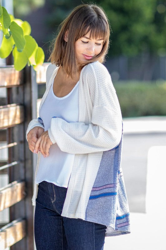 A woman leans against a wooden fence, looking over her shoulder. She is wearing the Plaid Back Waffle Knit Cardigan and jeans, embodying a cozy, effortless style.