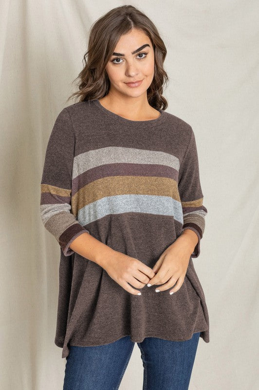 A person is standing against a beige backdrop, wearing a Plus Knit Stripe Panel A Line Tunic with horizontal stripes in various shades and blue jeans.