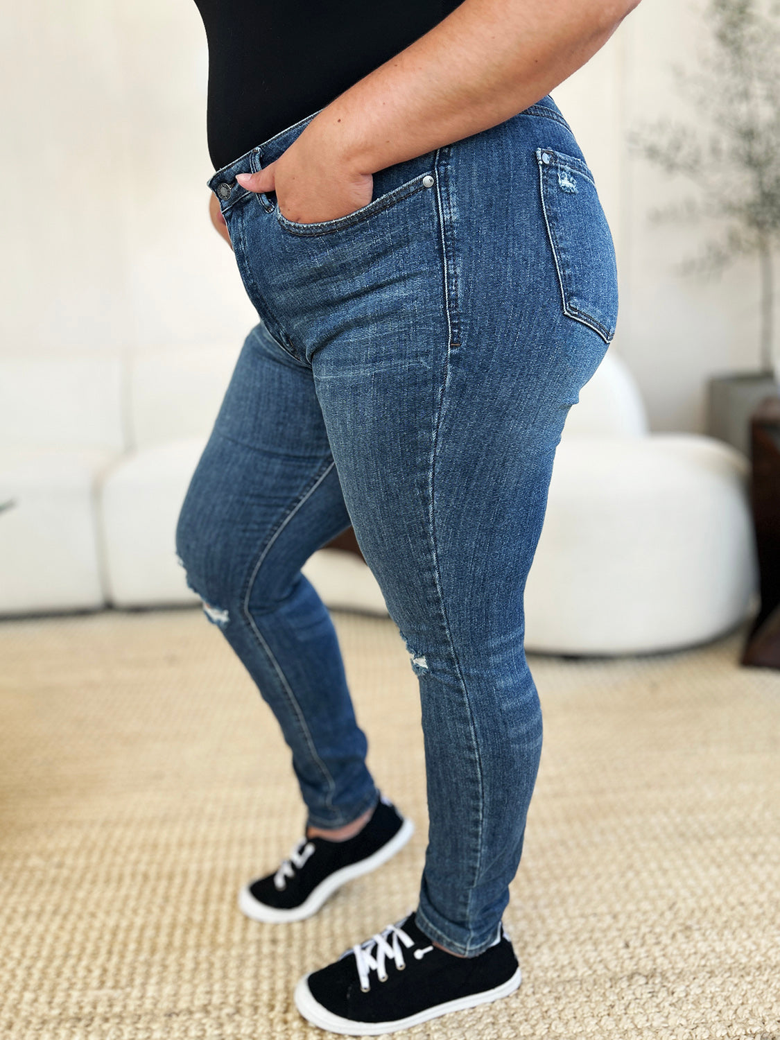 Person standing indoors wearing Judy Blue Full Size Mid Waist Distressed Slim Jeans, a black top, and black sneakers with white laces. There is a white sofa and a textured rug in the background.