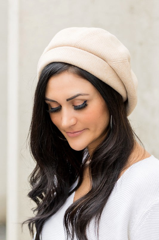 A smiling woman wearing a beige sweater and a Fold Over Beret leans against a stone wall.