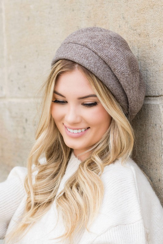 A smiling woman wearing a beige sweater and a Fold Over Beret leans against a stone wall.