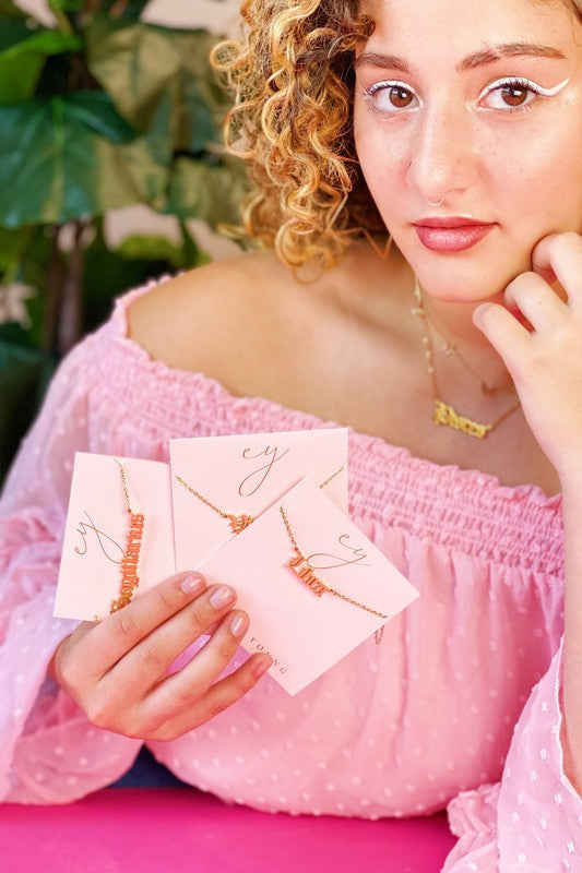 A person wearing a pink top holds three Old English Zodiac Necklaces, each on stainless steel gold-plated chains with pink note cards. The individual has curly blonde hair and white eyeliner, while a plant is visible in the background.