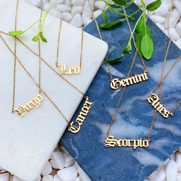 A person wearing a pink top holds three Old English Zodiac Necklaces, each on stainless steel gold-plated chains with pink note cards. The individual has curly blonde hair and white eyeliner, while a plant is visible in the background.