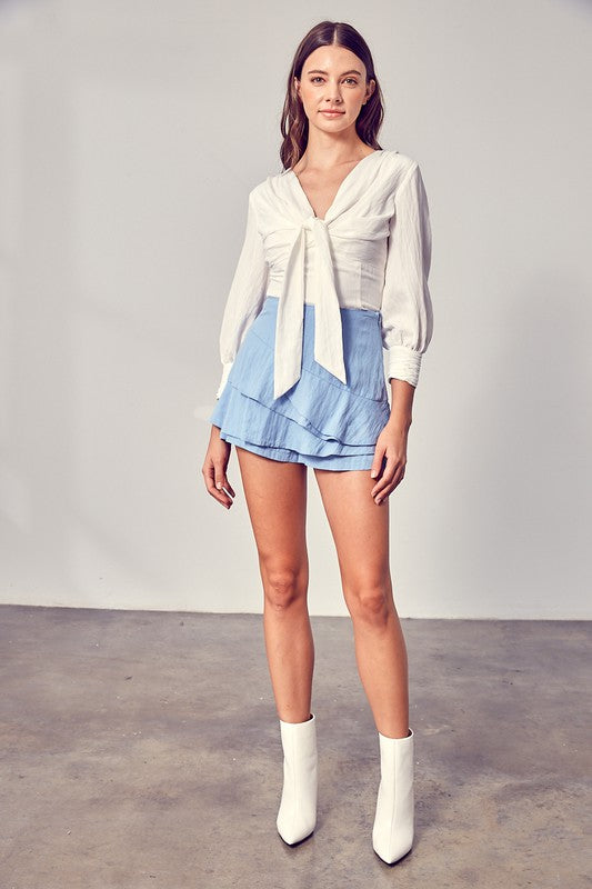 Against a neutral backdrop, a woman showcases elegance and comfort in a stylish Front Tie Long Sleeve Top, perfectly paired with a light blue layered skirt, making it a versatile wardrobe essential.