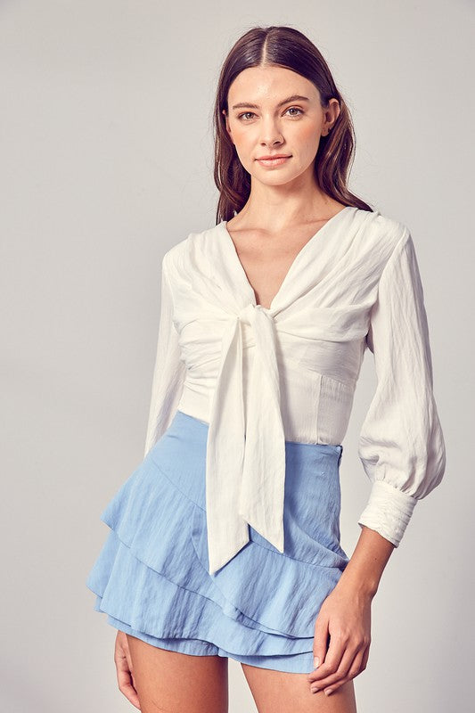 Against a neutral backdrop, a woman showcases elegance and comfort in a stylish Front Tie Long Sleeve Top, perfectly paired with a light blue layered skirt, making it a versatile wardrobe essential.