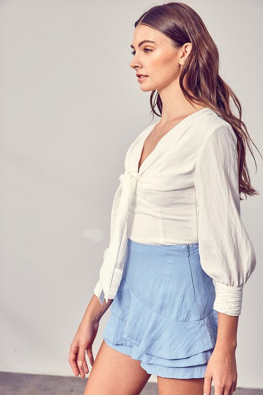 Against a neutral backdrop, a woman showcases elegance and comfort in a stylish Front Tie Long Sleeve Top, perfectly paired with a light blue layered skirt, making it a versatile wardrobe essential.