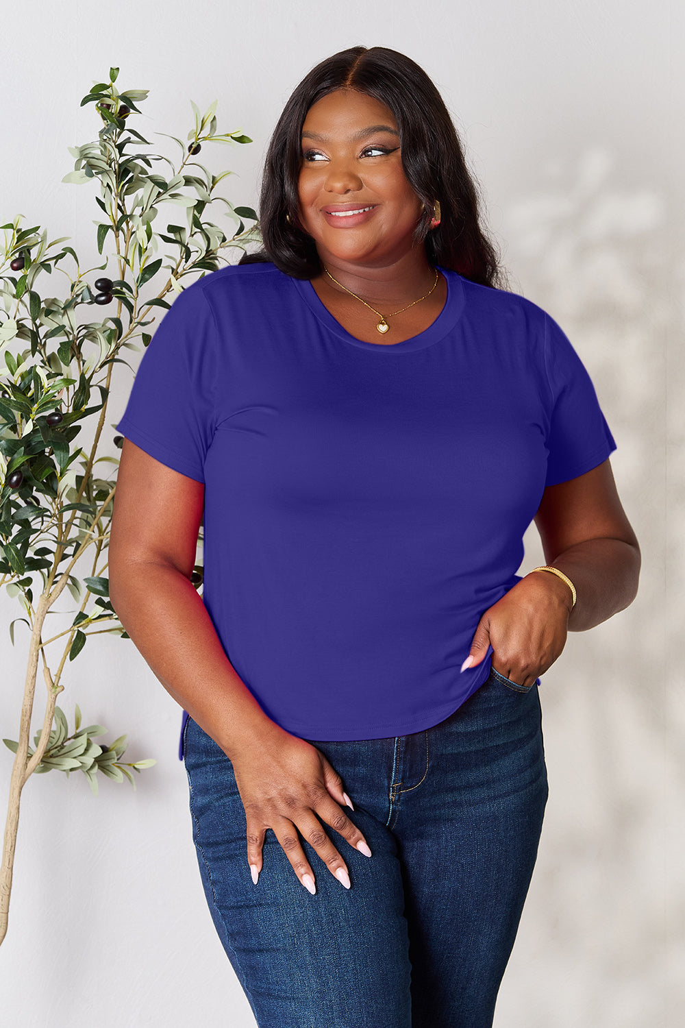 A woman with long hair, smiling with her right hand in her pocket and standing next to a plant, is wearing the Basic Bae Full Size Round Neck Short Sleeve T-Shirt in black along with beige pants made of rayon spandex.