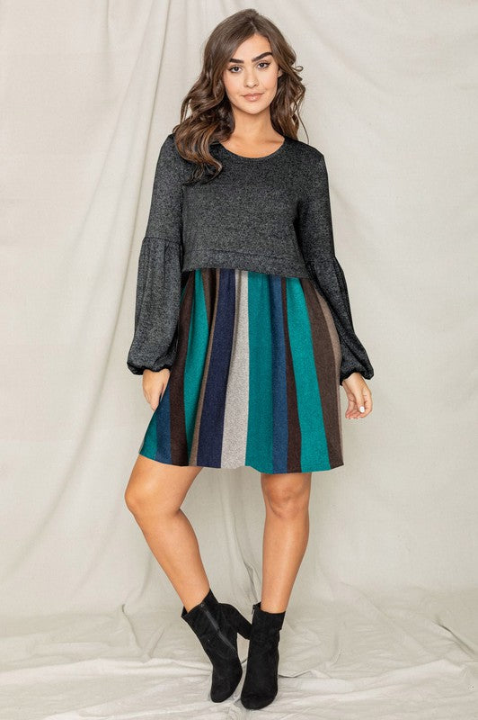 A woman wearing the Balloon Sleeve Pullover Dress, which features a black long-sleeve top paired with a multi-colored striped skirt made from Polyester Spandex. She has her left hand in the pocket of her skirt and is looking to her left.