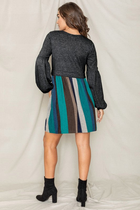 A woman wearing the Balloon Sleeve Pullover Dress, which features a black long-sleeve top paired with a multi-colored striped skirt made from Polyester Spandex. She has her left hand in the pocket of her skirt and is looking to her left.