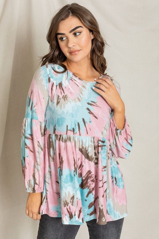 A woman with wavy brown hair smiles while wearing a loose-fitting Swirl Tie Dye Bishop Sleeve Tunic in shades of pink, blue, and brown. She stands against a neutral backdrop.