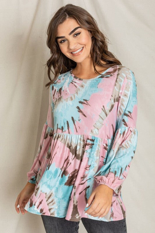 A woman with wavy brown hair smiles while wearing a loose-fitting Swirl Tie Dye Bishop Sleeve Tunic in shades of pink, blue, and brown. She stands against a neutral backdrop.