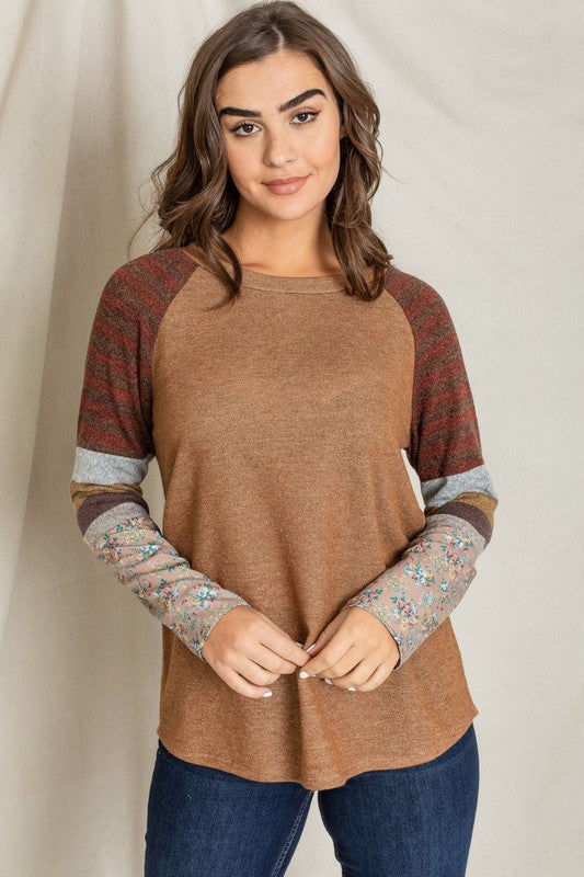 A woman stands against a plain background, wearing a Color Block Sleeve Tunic with brown body and multicolored striped and floral-patterned sleeves. Made in the United States from a comfortable polyester cotton blend, she rests her right hand on her neck and looks at the camera.