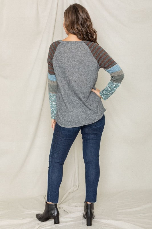 A dark-haired woman in a long-sleeved, multi-patterned Color Block Sleeve Tunic made from a polyester-cotton blend and jeans is smiling with her left arm raised, standing against a plain background.