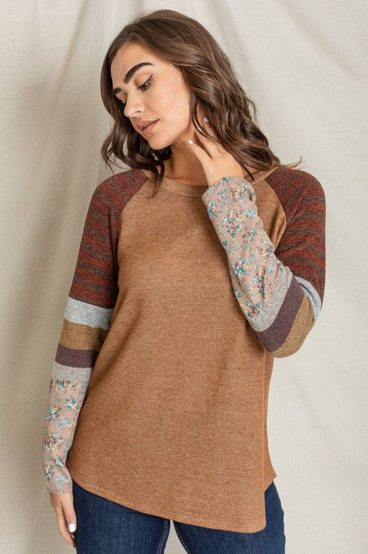A woman stands against a plain background, wearing a Color Block Sleeve Tunic with brown body and multicolored striped and floral-patterned sleeves. Made in the United States from a comfortable polyester cotton blend, she rests her right hand on her neck and looks at the camera.
