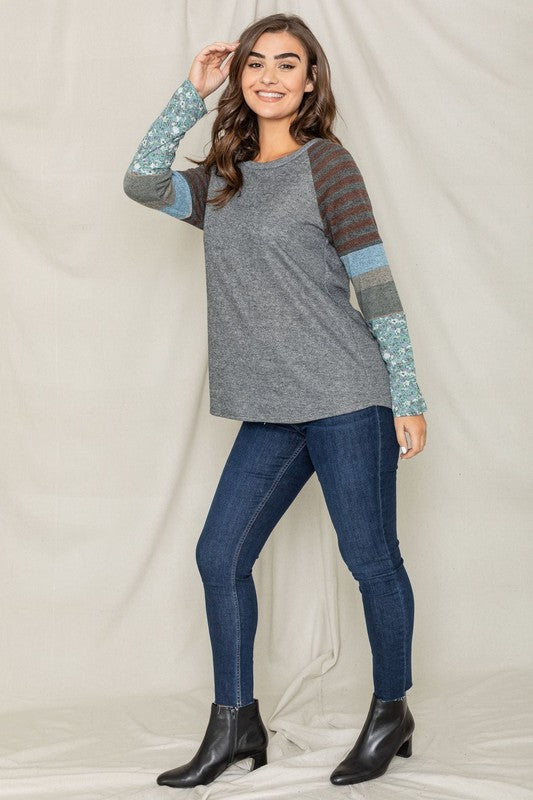 A dark-haired woman in a long-sleeved, multi-patterned Color Block Sleeve Tunic made from a polyester-cotton blend and jeans is smiling with her left arm raised, standing against a plain background.