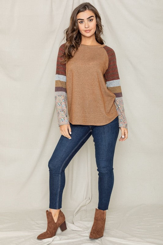 A dark-haired woman in a long-sleeved, multi-patterned Color Block Sleeve Tunic made from a polyester-cotton blend and jeans is smiling with her left arm raised, standing against a plain background.