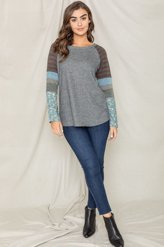 A dark-haired woman in a long-sleeved, multi-patterned Color Block Sleeve Tunic made from a polyester-cotton blend and jeans is smiling with her left arm raised, standing against a plain background.