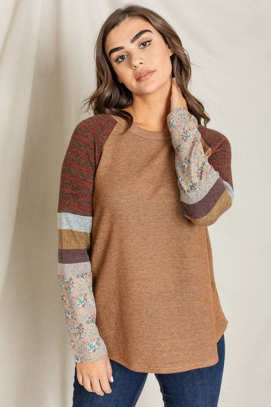 A woman stands against a plain background, wearing a Color Block Sleeve Tunic with brown body and multicolored striped and floral-patterned sleeves. Made in the United States from a comfortable polyester cotton blend, she rests her right hand on her neck and looks at the camera.