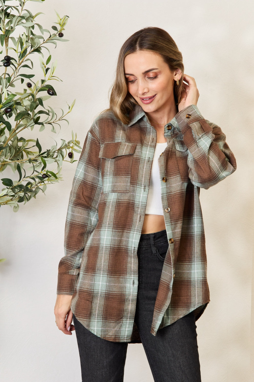 A woman stands in front of a light-colored wall and an olive tree, wearing the Mandy Plaid Dropped Shoulder Shirt paired with a white crop top and dark jeans. She smiles and holds her hair back with one hand.